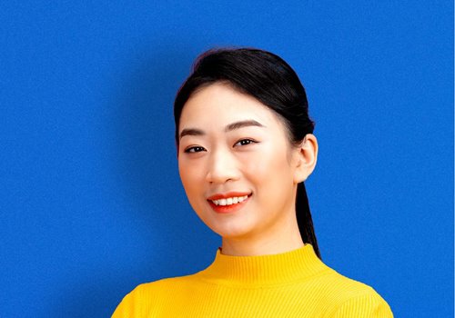A girl in a yellow sweater stands in front of a blue background smiling