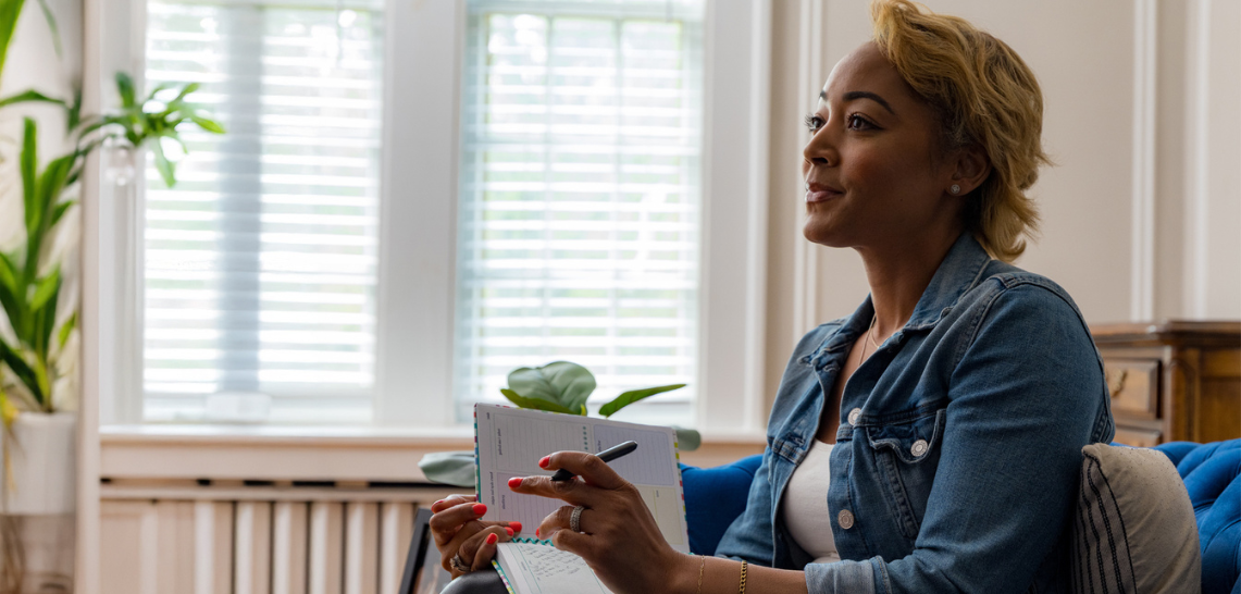Woman holding notebook and pen, sitting by a bright window.